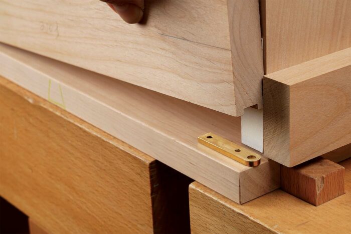 Mortise-location trick. Because the case top and bottom overhang the door, the fence must come off the jig for routing the hinge mortises in the case. To locate those, Gochnour mortises a setup block, sticks the hinge into the mortise with double-stick tape on top, and slides the block down into the door position, leaving the hinge in just the right spot. Then he pencils around it to guide the template location for routing.